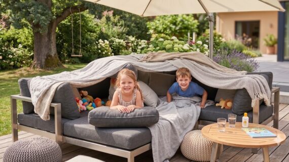Ein Junge und ein Mädchen schauen stolz aus ihrer Höhle, die sie auf einem Loungesofa auf der Terrasse gebaut haben (Bild KI-generiert)